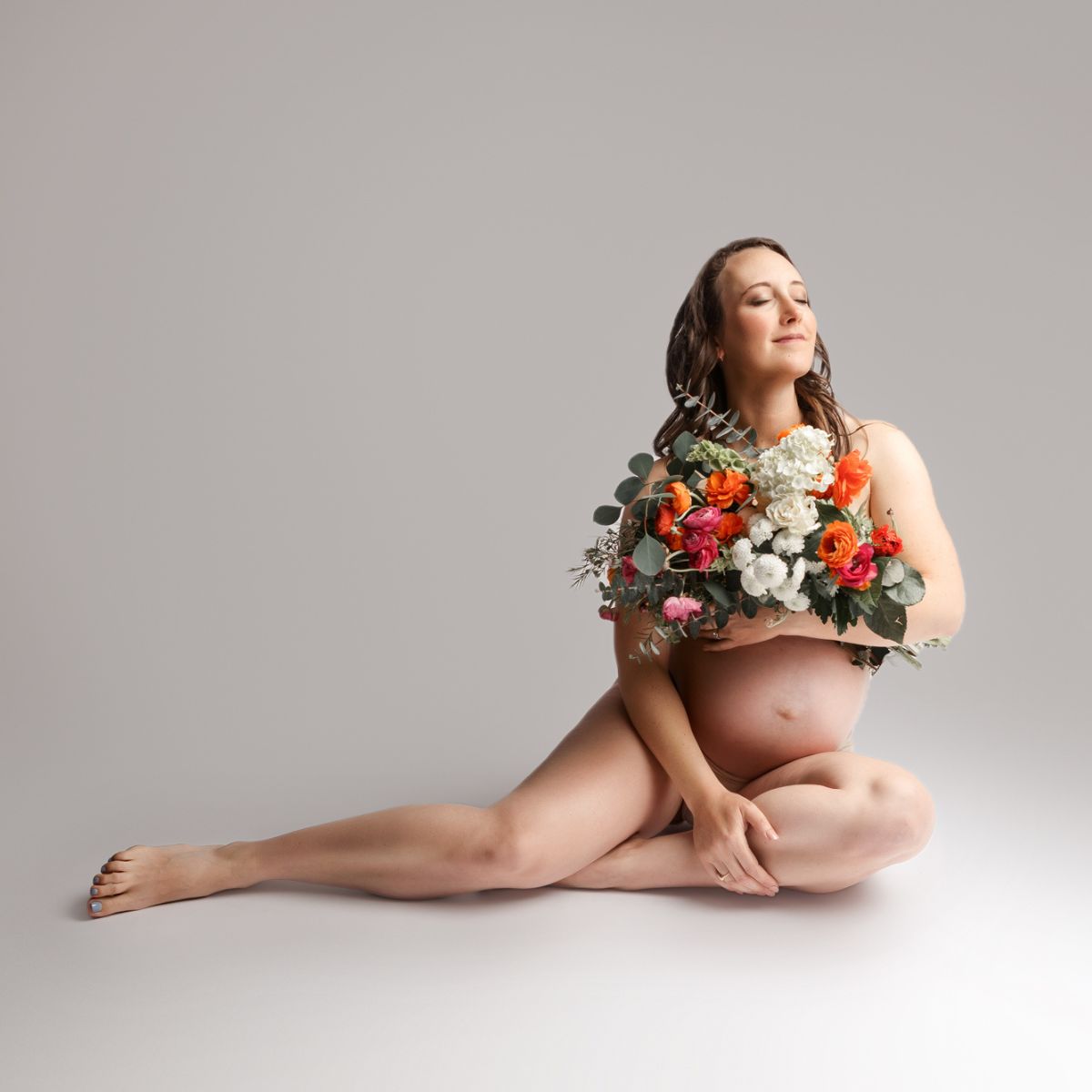 Pregnant woman seated on a white studio floor holding a bright bouquet of flowers over her chest