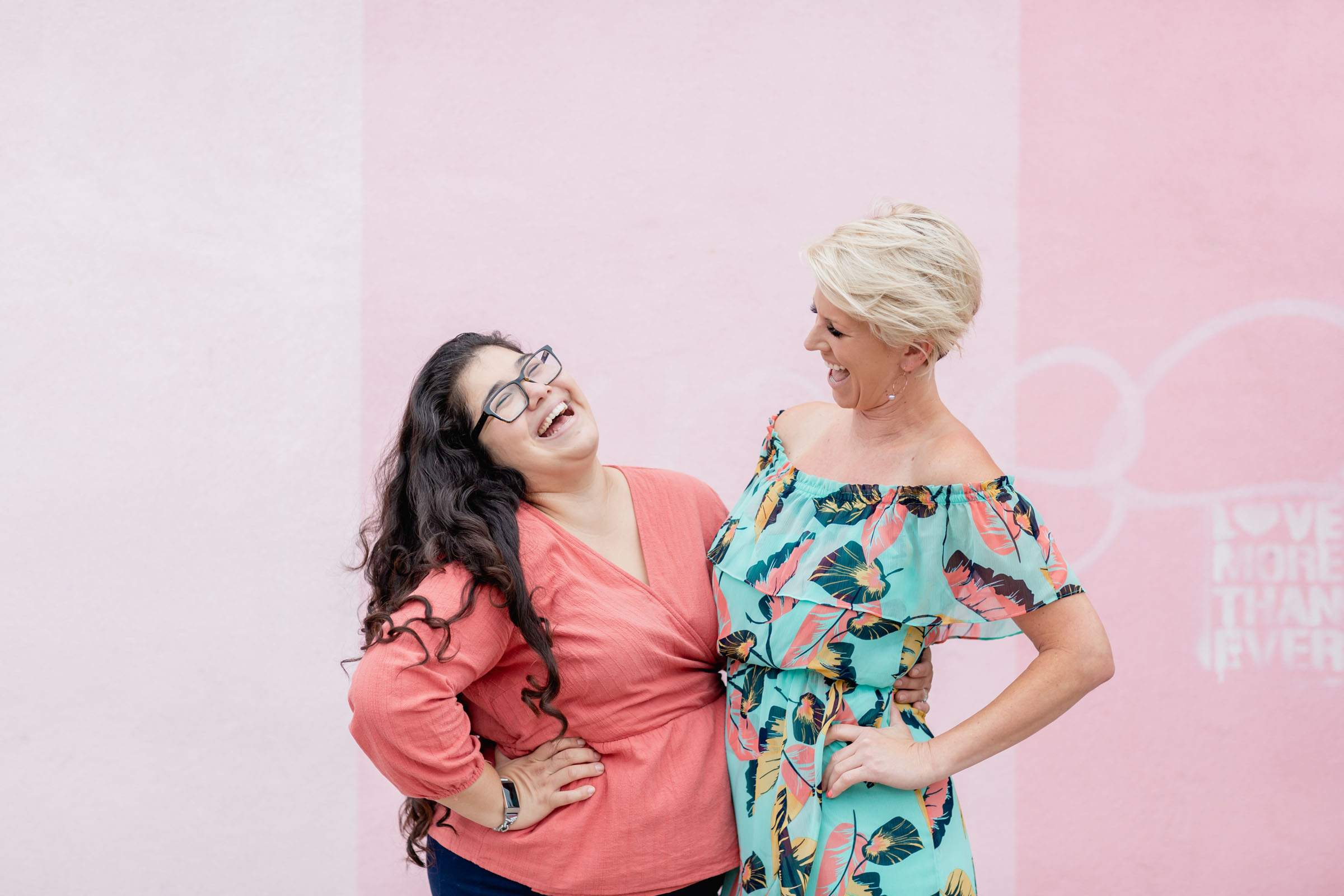 Two moms laughing together in front of a pink wall