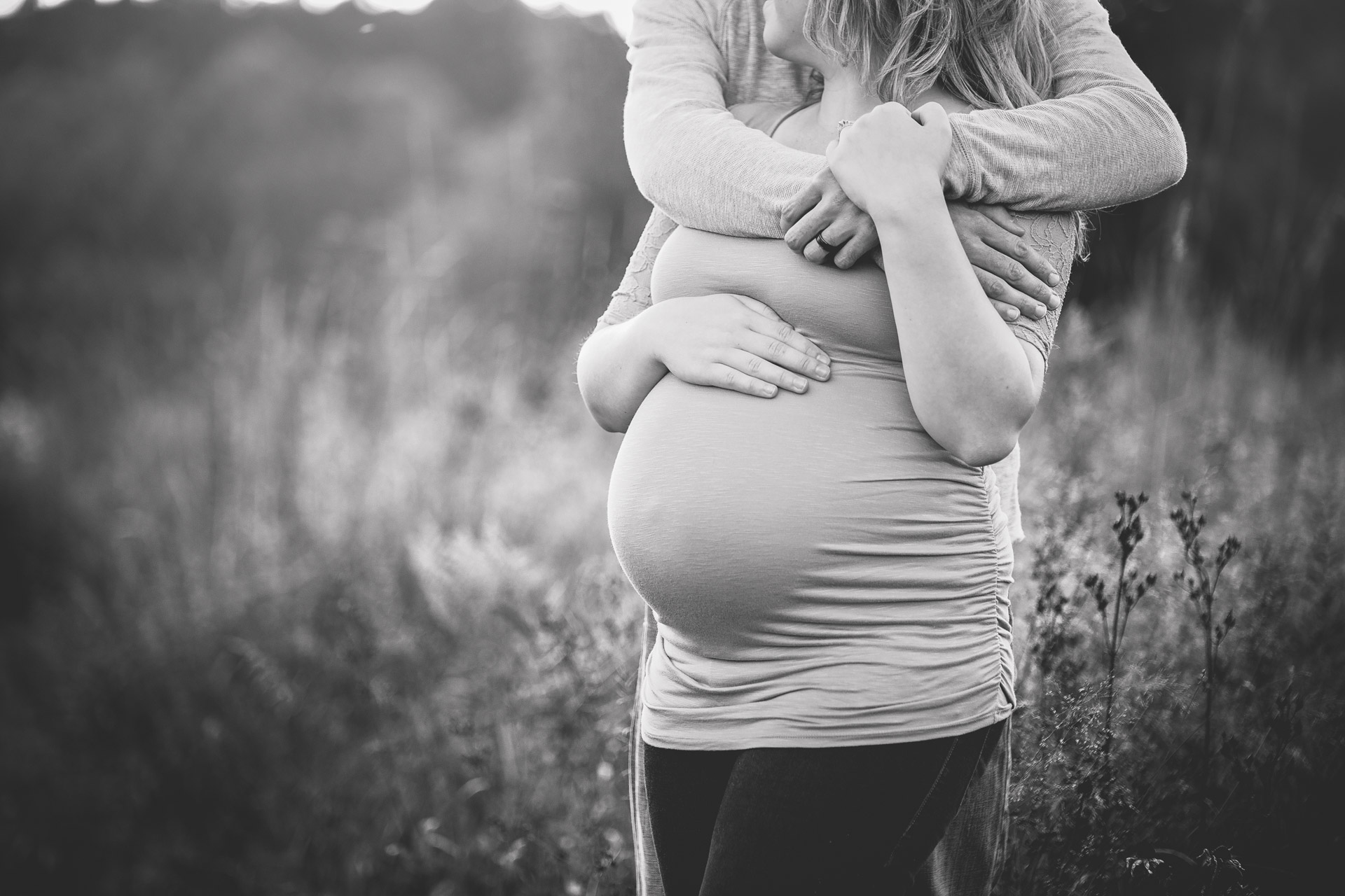 Black and white close-up of a couple's arms wrapped around her bump in a meadow