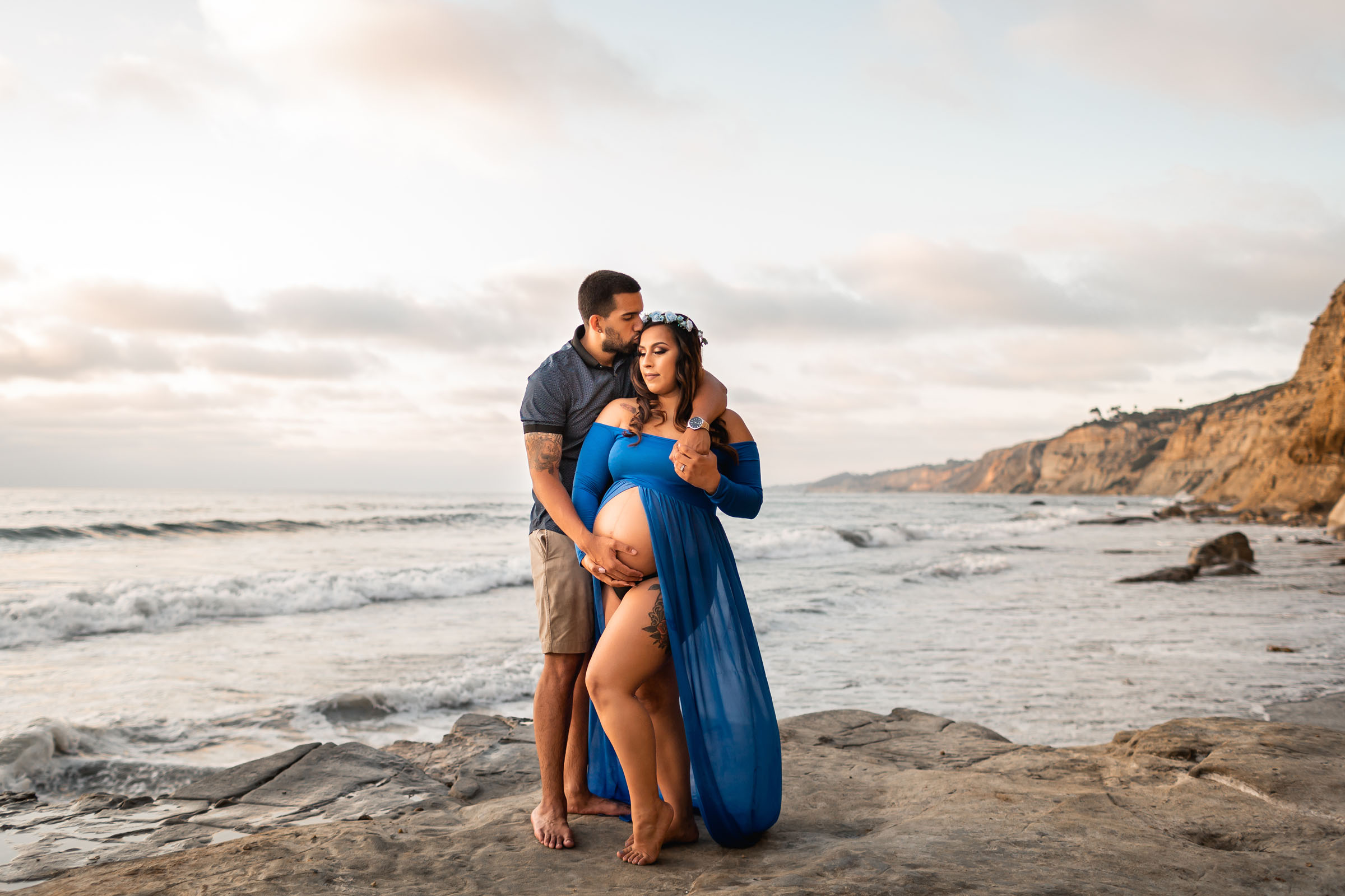 Expectant couple embracing on the rocks at sunset, she in a sheer royal blue gown
