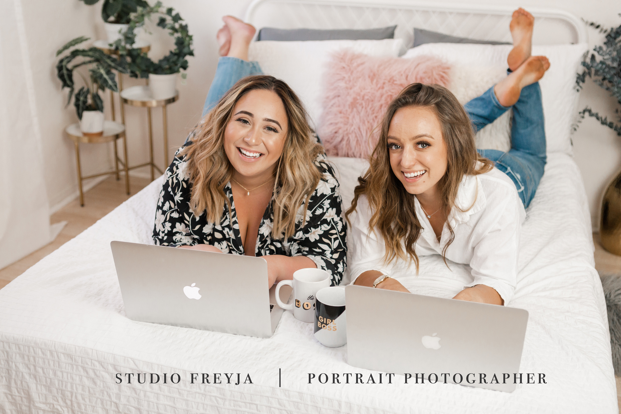 Two businesswomen lying on their stomachs on a bed with laptops, laughing at the camera