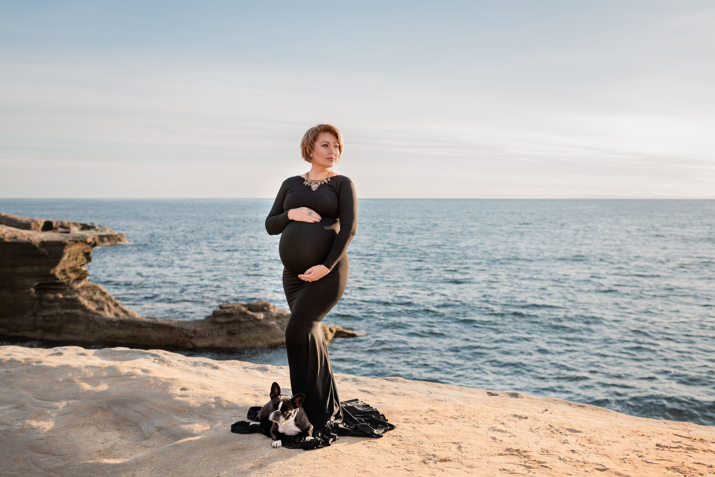 Pregnant woman in a fitted black gown standing on the sandstone cliffs at Sunset Cliffs