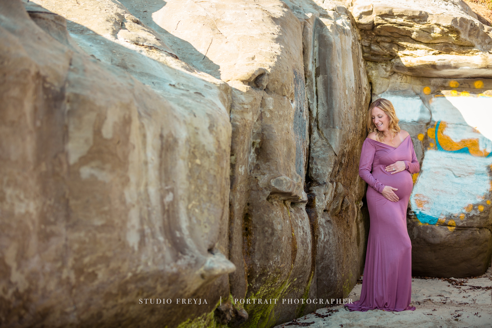 Pregnant woman in a mauve gown standing in a sandstone alcove on the beach