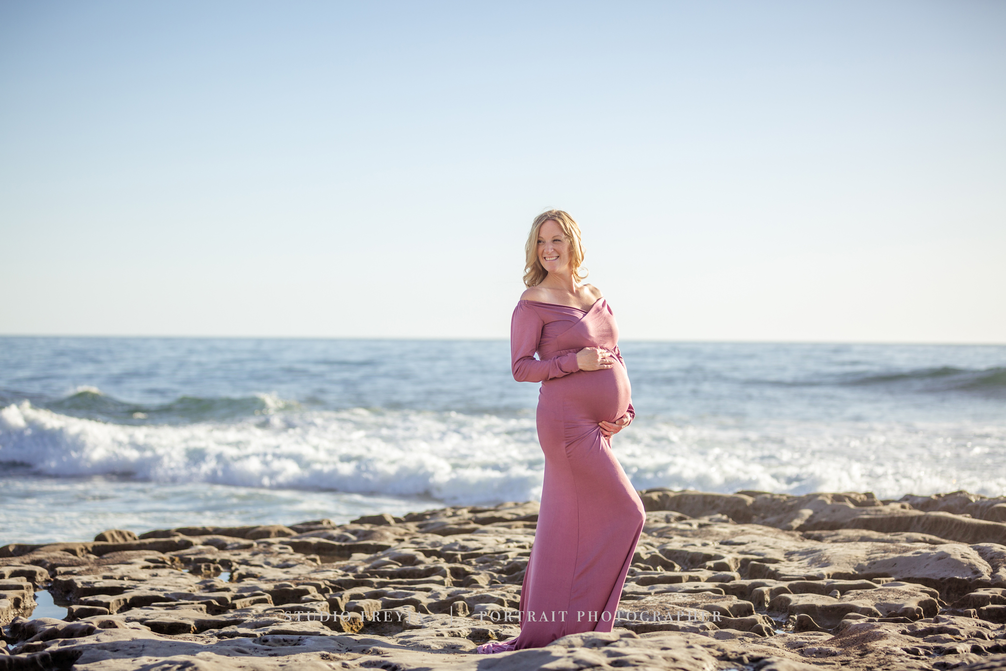 Pregnant woman in a mauve gown standing on the tide-pool rocks with waves behind her