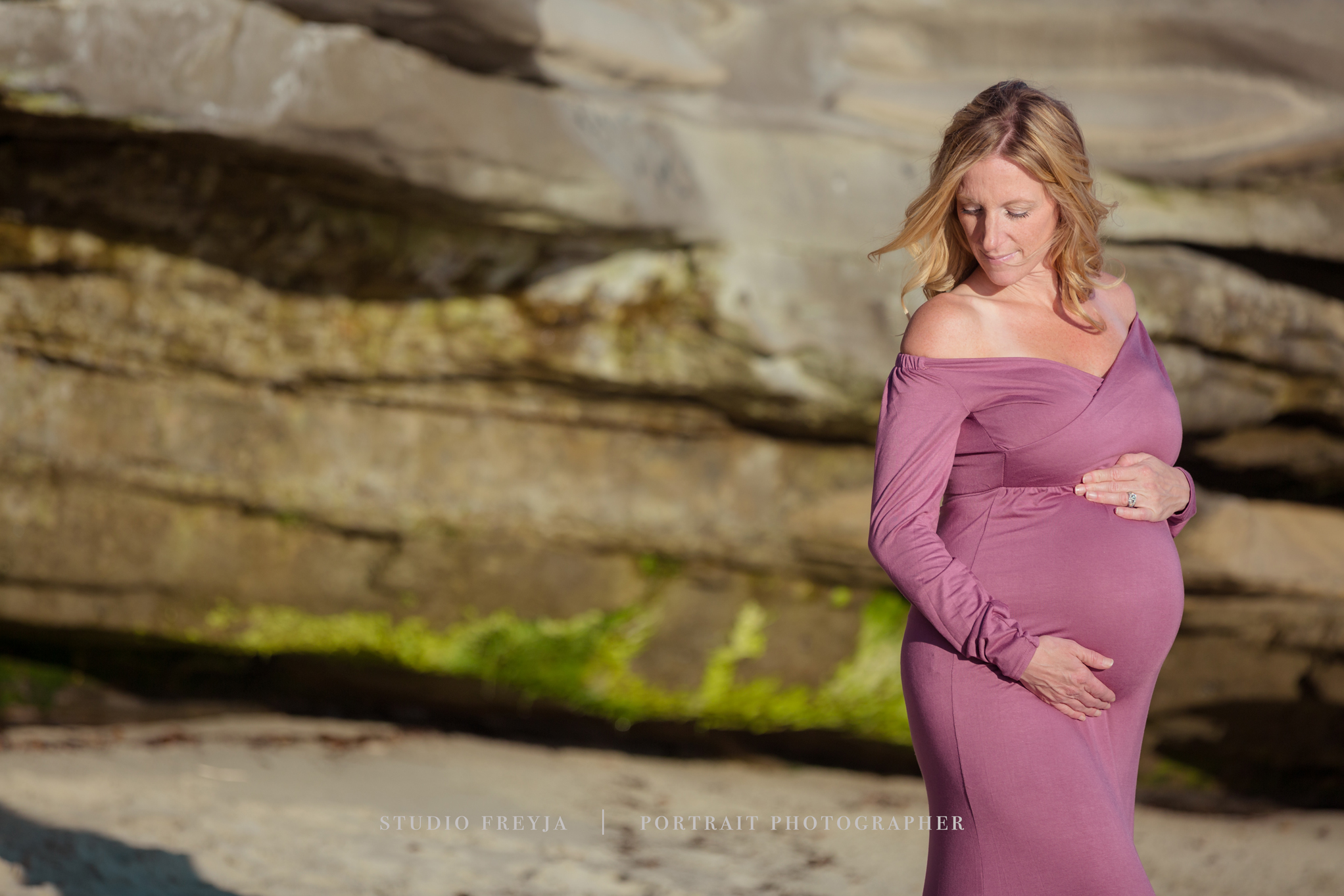 Pregnant woman in a mauve gown looking down at her bump in front of layered sandstone