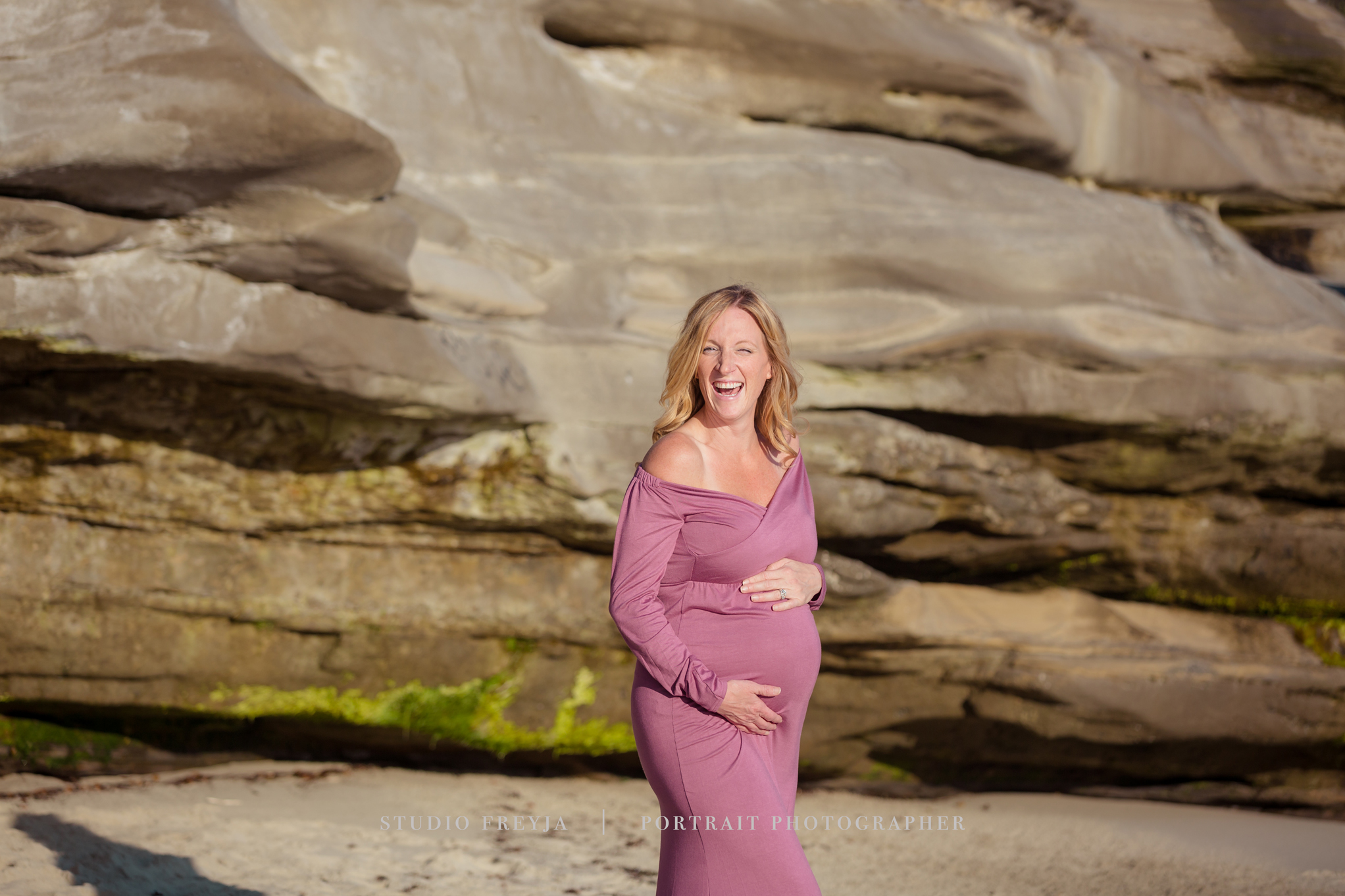 Laughing pregnant woman in a mauve gown on the beach in front of sandstone cliffs