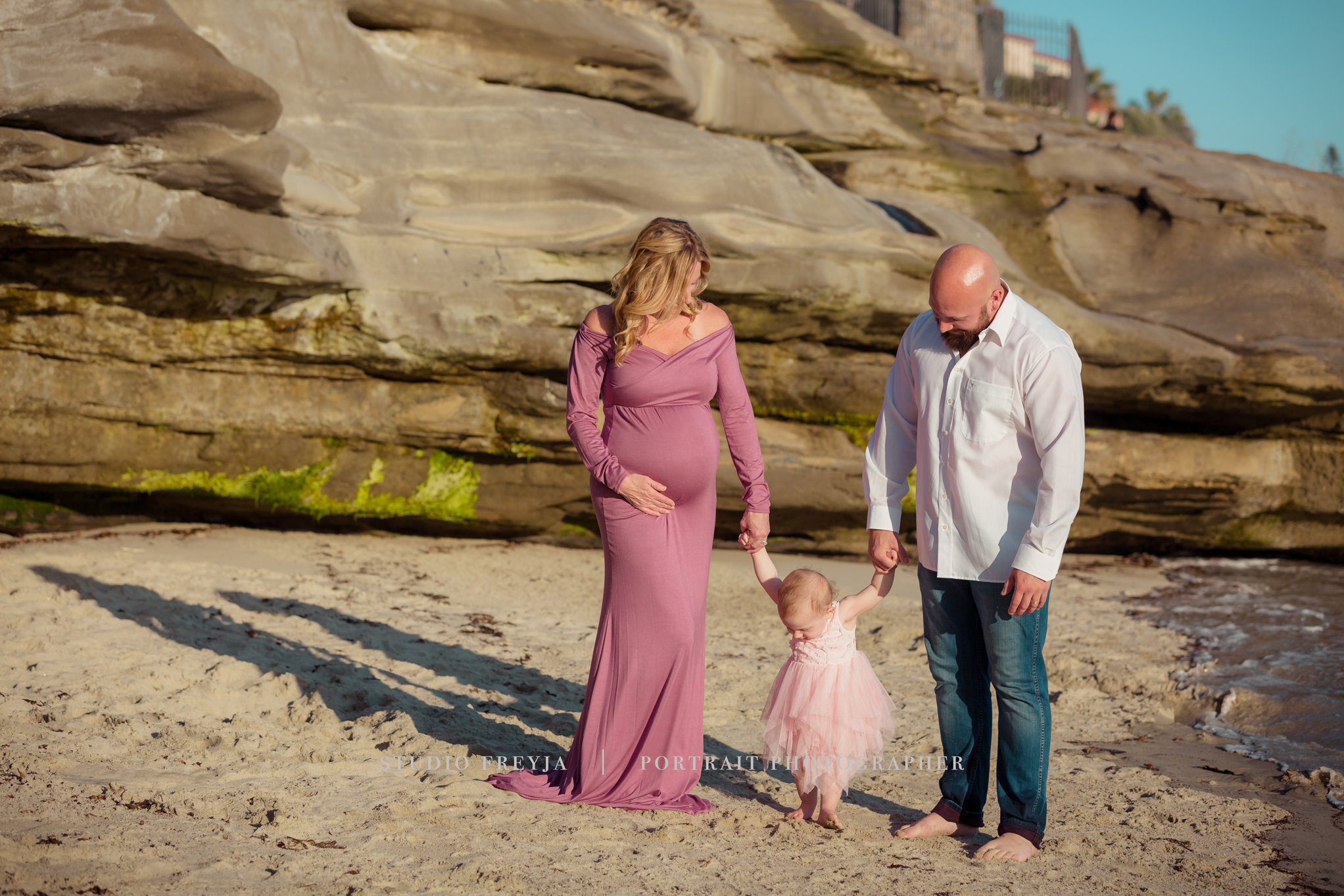 Toddler in a pink tutu holding her dad's hand and walking toward her pregnant mom on the beach
