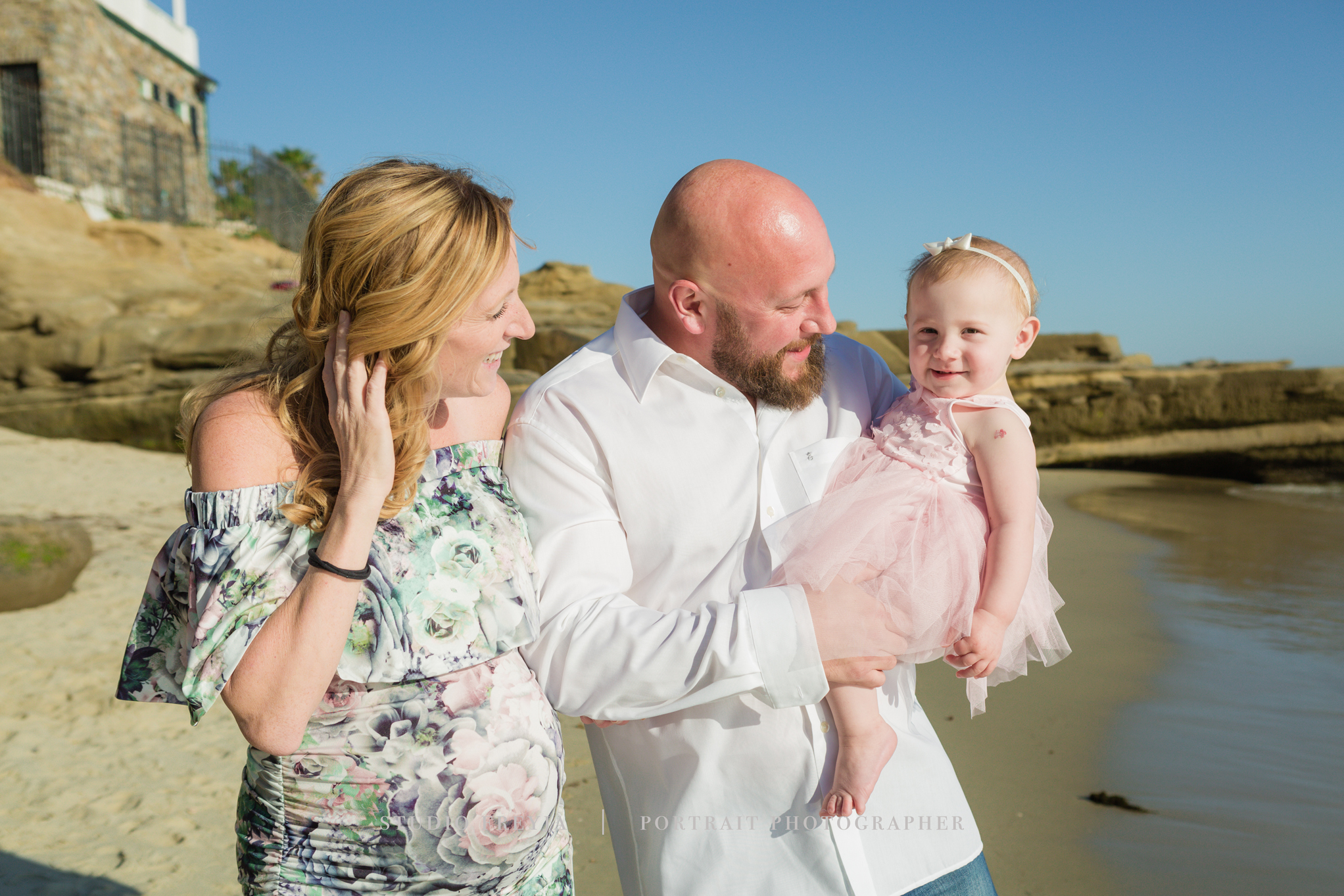 Expectant parents laughing with their toddler daughter on a La Jolla beach